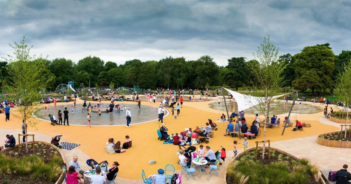 The paddling pools and playground in Cassiobury Park on a busy day, families spread across the splash area with mature trees behind