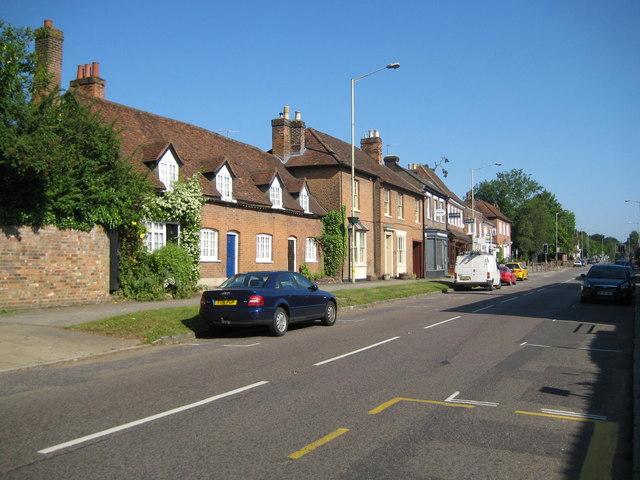 Old brick cottages fronting directly onto the main road through Kings Langley, with a car pulled onto the verge and the road running on past the shops