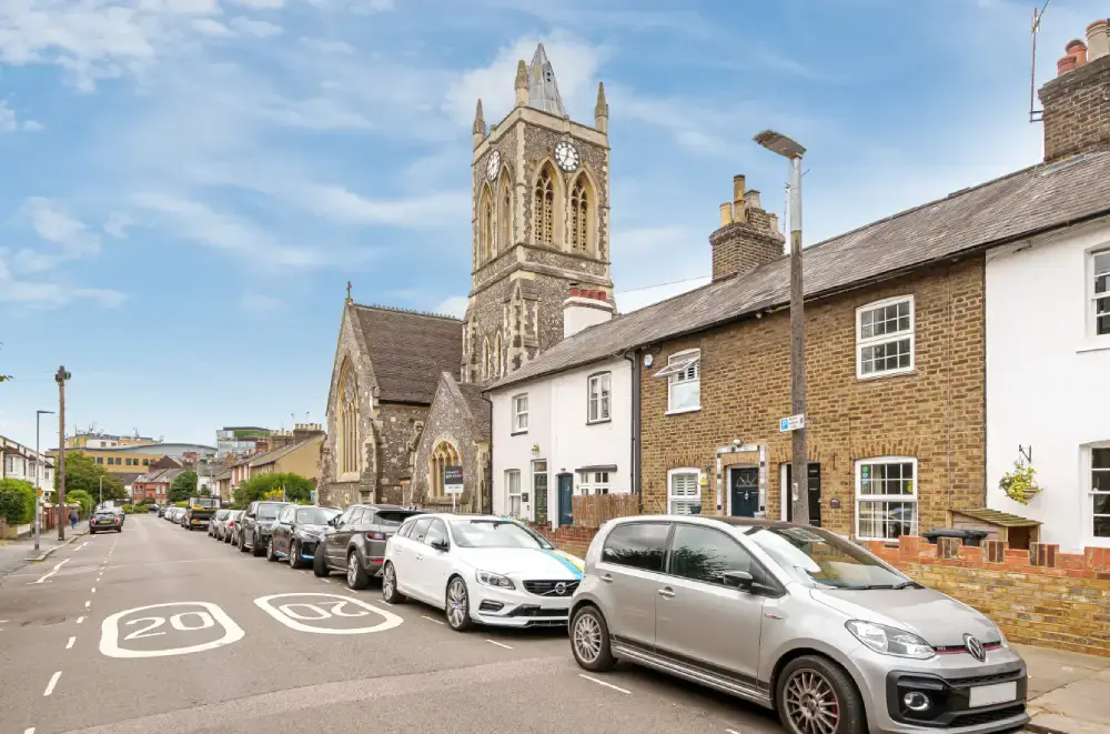 A residential street in Nascot Wood, Watford, parked nose to tail on both sides, with brick cottages fronting straight onto the pavement and a flint church tower behind